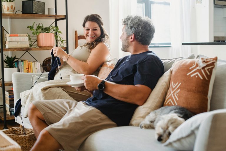 Side view of crop adult man in home clothes sitting on couch with hot drink and looking at smiling pregnant woman knitting in cozy apartment