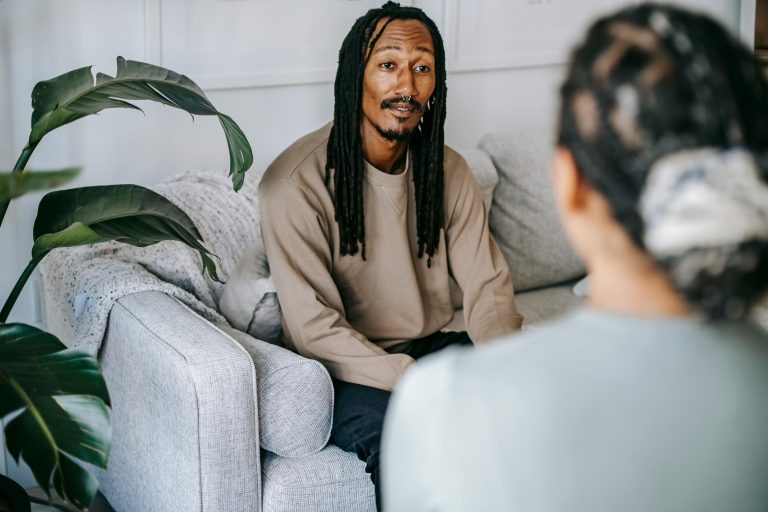 Content African American male with dreadlocks wearing casual outfit having conversation with female friend while sitting on couch in cozy living room